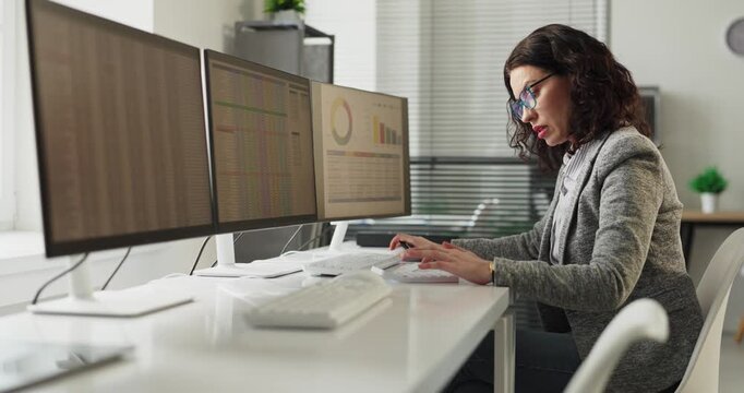 Woman analyst studies data charts at computer. Focused professional in an office reviews graphs across three monitors for finance reporting and work insights. Clear data driven business concept.
