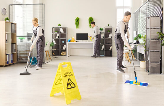 Cleaners janitors team mopping office for sanitation. Uniformed crew vacuuming floors and wipe desks as a wet floor sign promotes safety in a bright workspace. Clean office safety sign concept.
