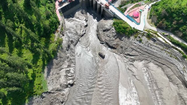 Cinematic aerial glide looking down at water control sluices and silt-choked riverbed at Barshaini dam, documenting renewable energy development in the Himalayas.