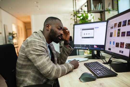 Stressed office worker rubbing forehead at dual monitors in modern office