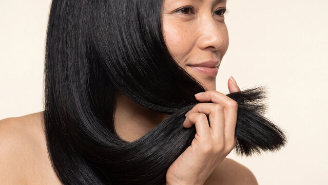 Woman holding a strand of her long straight black hair to showcase its healthy texture and shine during a close up beauty care product commercial shoot.
