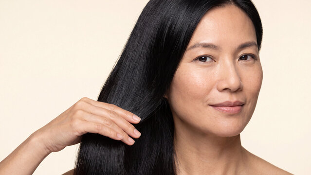 Mature woman gently touches her long glossy black hair with a serene expression while displaying healthy and well-maintained strands against a neutral plain background.