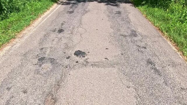 Close view of a worn asphalt road with visible cracks and patches, camera moving steadily forward, shifting tree shadows crossing the cracked gray surface in bright daylight through the forest.