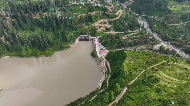 High angle drone tracking shot over the Parbati-2 HEP Dam and sediment-heavy reservoir in Barshaini, a key energy infrastructure site in Parvati Valley, Himachal Pradesh.