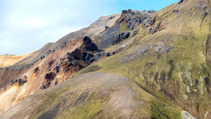 Obraz premium Colorful layered mountains with moss-covered slopes and dark rock formations create a dramatic landscape in Landmannalaugar, Icelandic highlands.