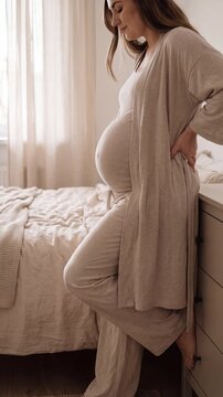 Pregnant woman standing in a sunlit bedroom, wearing comfortable beige loungewear and holding her belly. The atmosphere is serene and peaceful. Vertical video made using Generative AI.