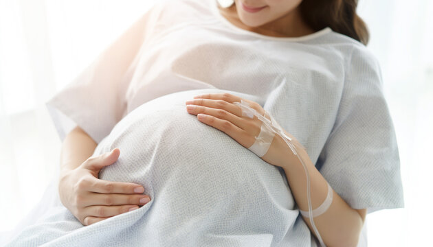 A pregnant woman rests on a bed while gently holding her baby bump with both hands in a bright indoor setting.