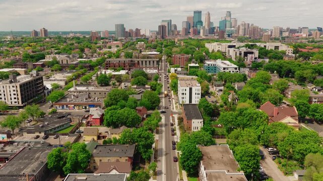 Aerial over a mixed use urban avenue in Minneapolis, Minnesota, with ground floor storefronts, new midrise housing, brick warehouses and green trees toward downtown.
