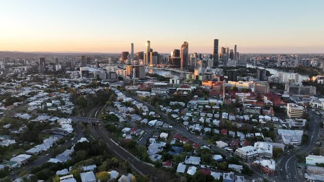 Slow forward tracking aerial shot over St Lucia suburbs toward the Brisbane CBD skyline, rooftops and treetops in the foreground, golden hour light, St Lucia, Brisbane, Queensland, Australia.