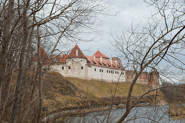 Bauska, Latvia - April 5, 2026: An ancient castle in the town of Bauska against a gray sky, with trees and a river in the foreground © fotofotofoto