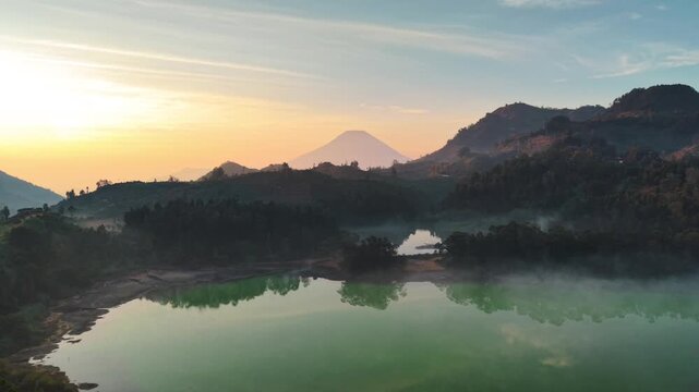 Stunning aerial view of a misty volcanic lake with green sulfur water surrounded by dense forest at sunrise, featuring rolling hills and a mountain peak in the background under soft golden light.
