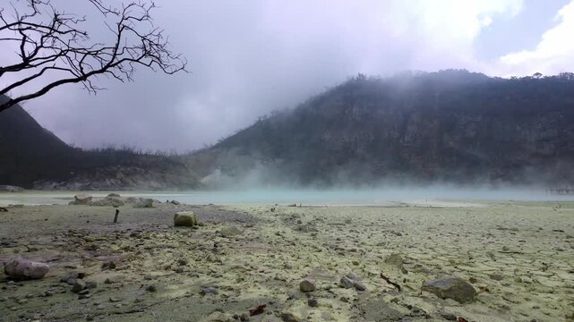 Ground-level static video of sulfur steam and thick mist slowly rising over the turquoise crater lake at Kawah Putih, surrounded by rocky terrain and volcanic hills in Ciwidey, Bandung, Indonesia