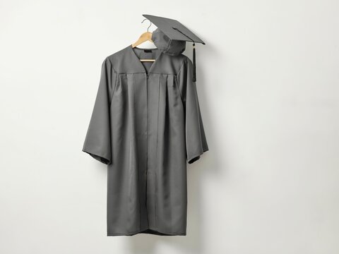 A grey graduation gown and a matching mortarboard cap hanging together on a wooden hanger against a neutral white background, representing academic success and upcoming commencement ceremonies