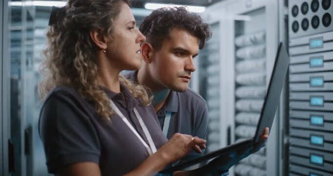 Focused Female System Administrator Types on Laptop While Colleague Conducts Server Cabinet Check in Data Center Corridor. Women in STEM Drive Modern Enterprise IT Operations and Cloud Security.