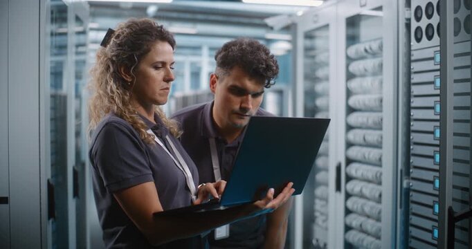 In Server Room Corridor, Two Network Engineers Inspect Cybersecurity Metrics and System Logs on Laptop Screen. Joint Diagnostics and Enterprise Cloud Monitoring Protect Critical Server Networks.
