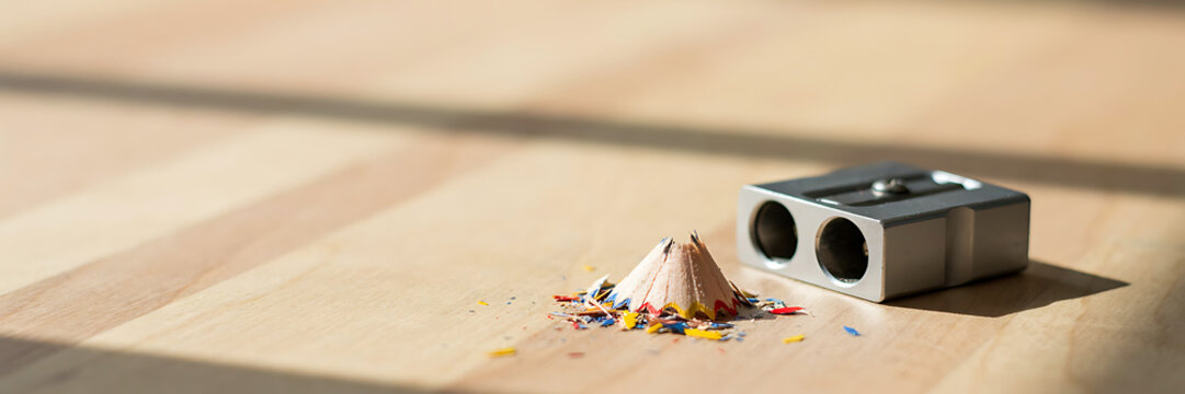 A metallic dual-hole pencil sharpener placed on a light wooden desk surface next to a small pile of colorful wood shavings from a pencil point