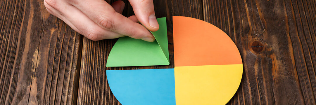 Human hand placing a green segment into a colorful paper pie chart on a dark brown rustic wooden table surface for business data analysis