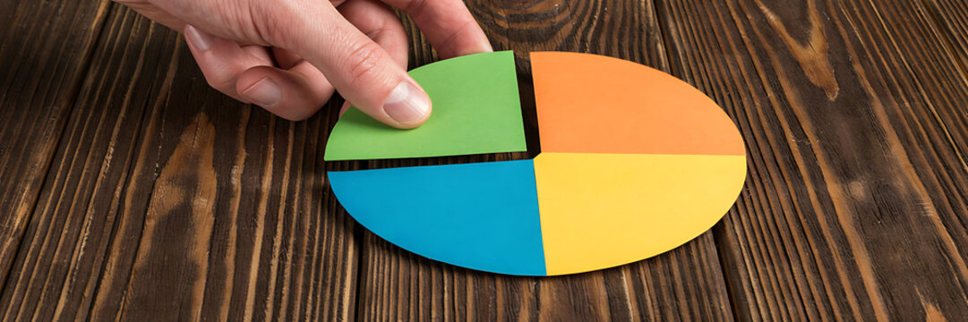 Human hand placing a green segment into a colorful paper pie chart on a dark brown rustic wooden table surface for business data analysis