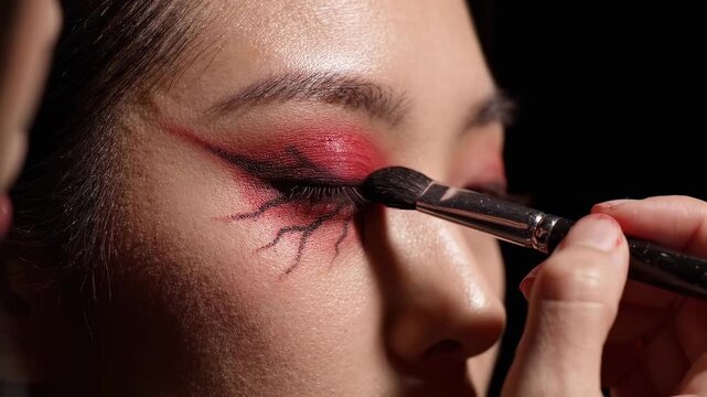 Close-up of a person's eye with dramatic red and black artistic makeup being applied by a brush.