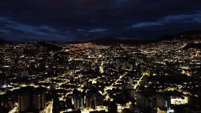 Wide Night Drone View of Quito Ecuador City Lights, Dense Urban Buildings, Mountain Horizon and Dramatic Cloudy Sky Over South American Capital