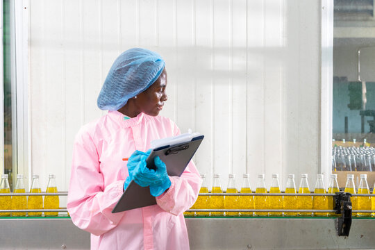 Black factory nutritious woman inspecting drink quality with rows of glass bottles filled with nutritious health drinks on the automated conveyor system in manufacturing assembly line.
