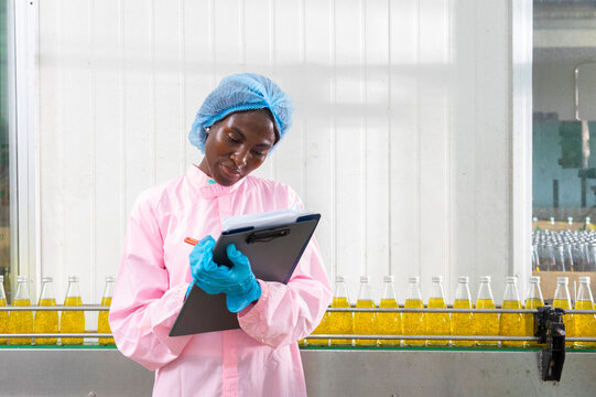 Black factory nutritious woman inspecting drink quality with rows of glass bottles filled with nutritious health drinks on the automated conveyor system in manufacturing assembly line.