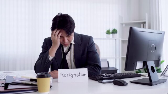 Stressed Asian businessman loosens tie holding resignation sign at office desk feeling burnout