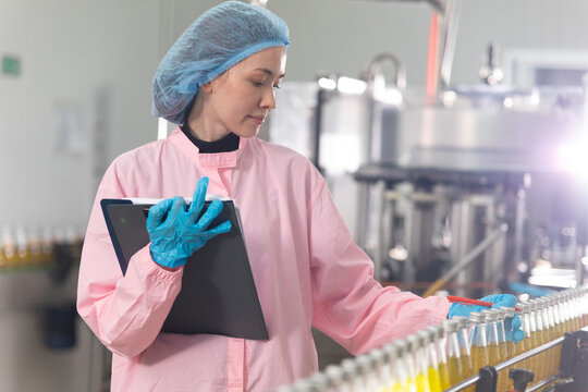 Young beautiful  white woman inspector standing in front of assembly line with capped bottled drinks and examining plastic packaging.
