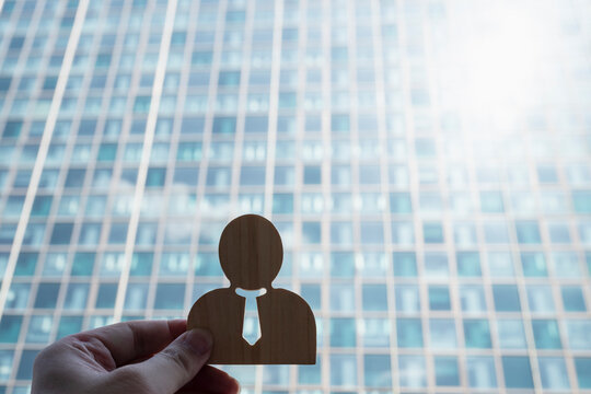 Executive recruitment and career promotion concept with hand holding wooden businessman figure against glass skyscraper office building with sunburst for corporate talent and leadership.