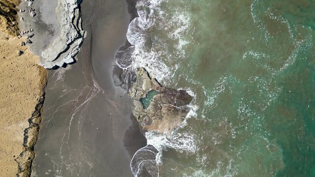Aerial view of waves washing over a rocky black sand beach, creating natural textures and patterns along the tropical coastline.
