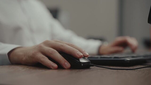 Closeup of male office worker hands typing on keyboard and using wired mouse at wooden desk. Business person performing daily tasks and data entry on personal computer in corporate setting.