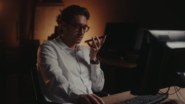 Professional male finishing voice message on mobile device and continuing to work late on desktop computer, dedicated to meeting deadline in dark office. Shooting in slow motion.