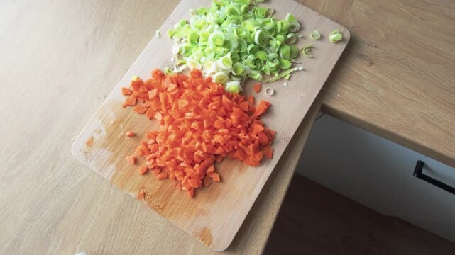 Detailed close-up shot of freshly chopped carrots and leeks arranged on a wooden cutting board, bathed in natural daylight on a modern wood table, perfect for cooking themes.