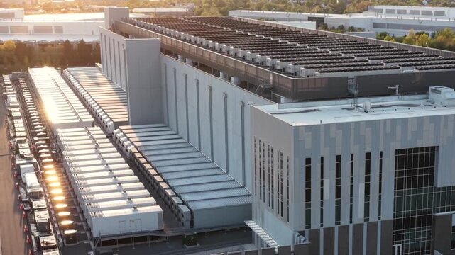 Backup generator enclosures line a data center hall in Ashburn, Virginia, golden hour sun catching the rooftop condensers and the trees along the service road. Aerial orbit shot