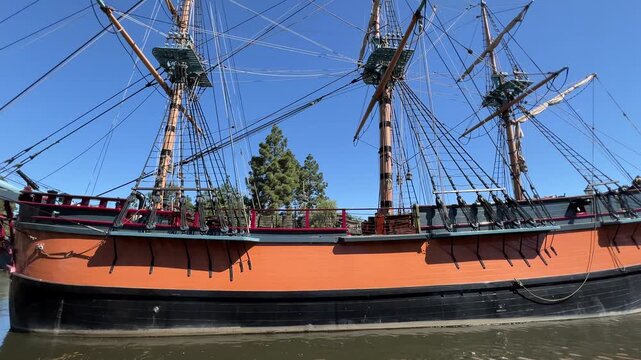 Orange and black wooden tall ship with rigging and masts slowly moving on calm water on a clear blue sky day, side profile tracking shot