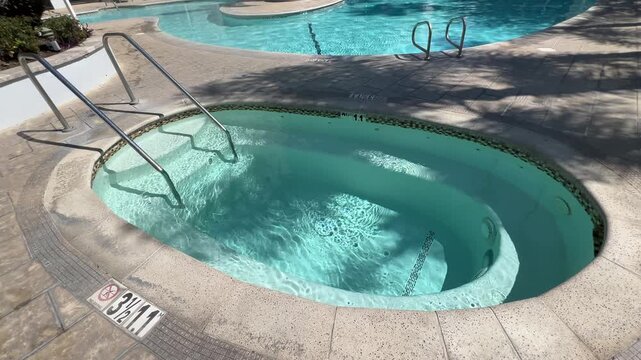Empty oval hot tub beside resort swimming pool in bright midday sun, bubbling turquoise water and clean pool deck