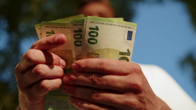 Closeup view of a young man counting 100 Euro banknotes outdoors. Guy flipping through a handful of 100 bills, money concept. Man counting cold hard cash money.