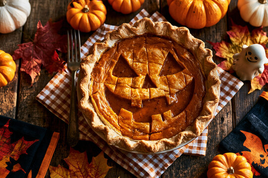 Pumpkin pie with jack-o-lantern face cut into crust, Halloween overhead flat lay with pumpkins and gingham napkin