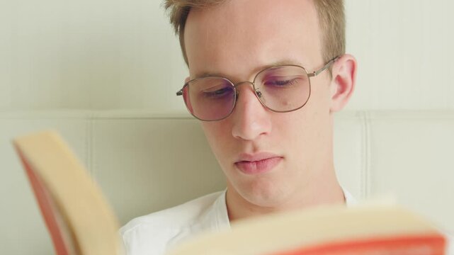Young student in eyeglasses reading a paperback book indoors. Portrait of a young man with glasses reading a book. Close-up of a student flipping pages in a book. Intelligent young man enjoying litera
