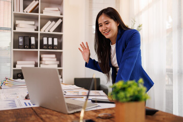 Professional businesswoman waving hello during a laptop video call, engaging in online business communication, remote work, or virtual meeting from a modern office setting © David