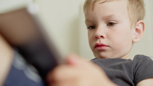 Close-up of a small boy focused on a digital tablet screen at home. Little child watching a cartoon on an iPad.