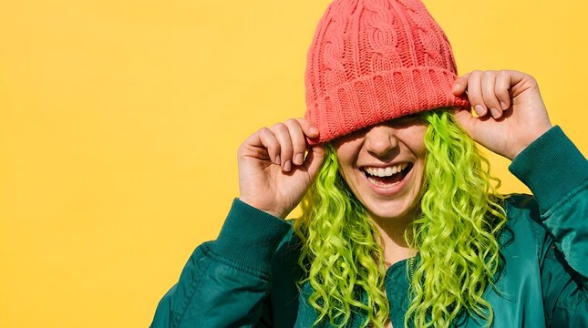 Cheerful woman with lime green hair wearing pink beanie over eyes on yellow background