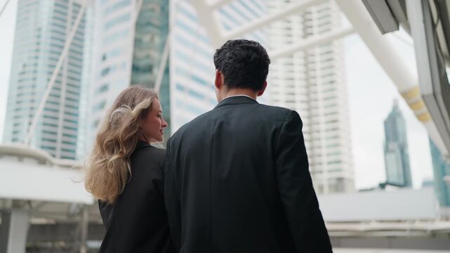 Two professionals walk together through a modern cityscape. They move side by side, focused forward with purpose. Tall buildings loom behind them