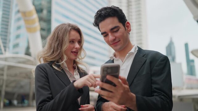 Two professionals share a moment outdoors. They smile, focused on their phones. Urban towers loom behind them. They converse while browsing content