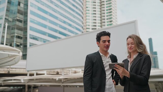 Business partners walk past a billboard in a city. They discuss ideas while holding tablets and gesturing. Their smiles suggest a positive