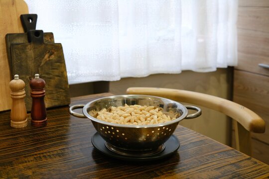 Draining soaked beans in colander on wooden kitchen table. Salt and pepper shakers are nearby. Sunlight filters through curtain. Kitchen space is organized and clean