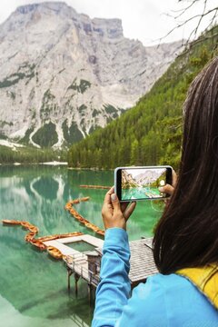 Portrait of Woman hiker taking smartphone photo of Lago di Braies alpine lake turquoise water mountain reflections Dolomites Italy