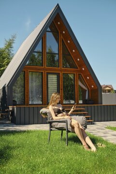 Slender woman lounging in a chair, enjoying the sun with a smartphone in her hand. She is scrolling the feed of social network and sharing her emotions