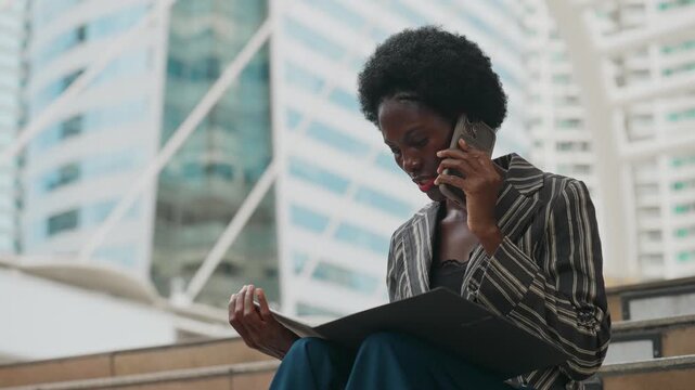 Focused professional talks on phone while reviewing documents. Urban setting with modern glass buildings in background