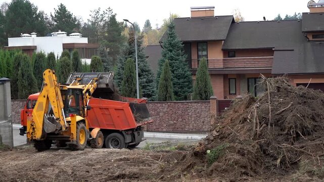 Yellow loader scoops earth beside a pile of branches. Brown house looms in background, surrounded by trees. Heavy machinery clears debris for new construction. , no animals, no plants identified